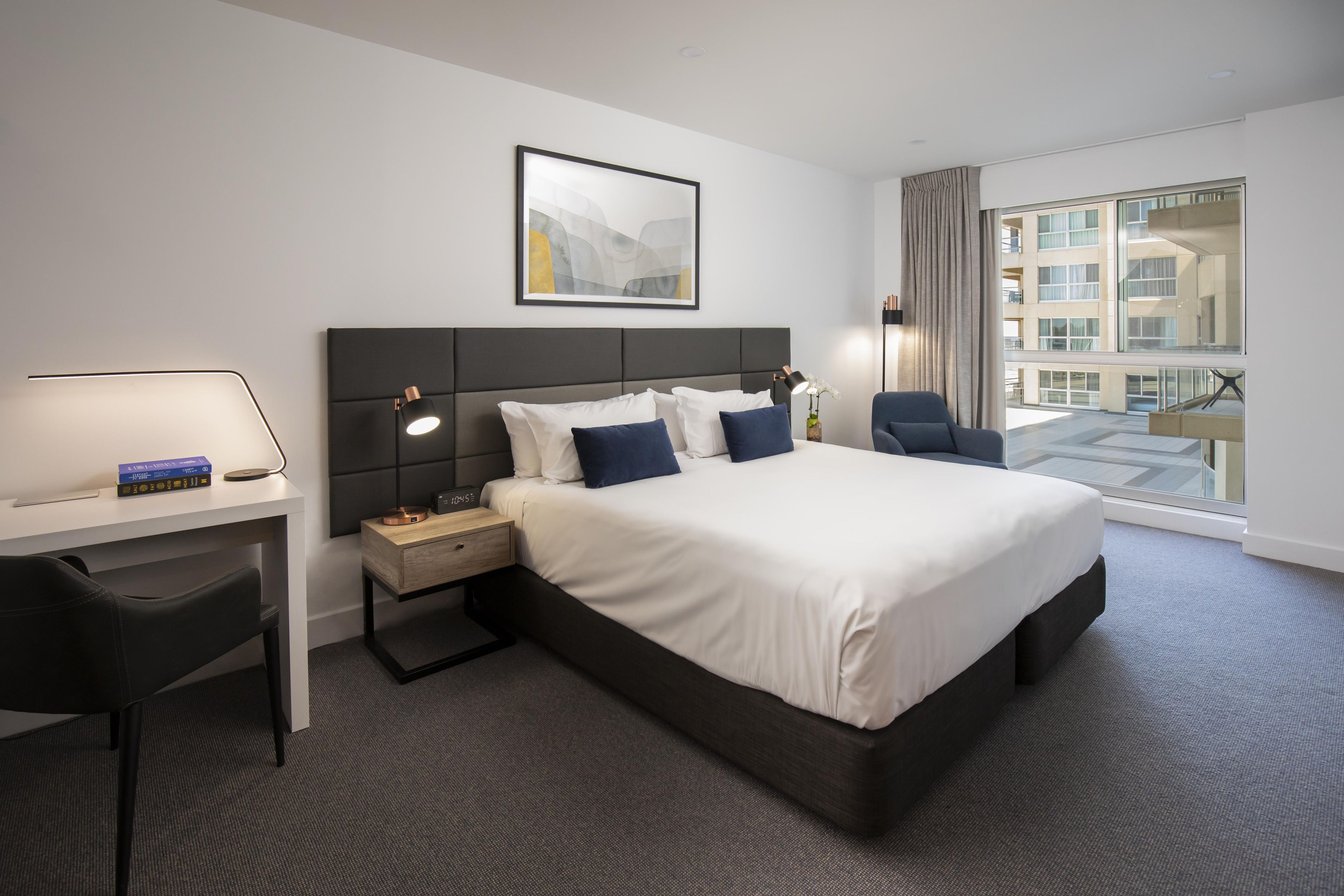 A hotel room with a large bed, a desk with a lamp, a chair, and a view of a building through the window.