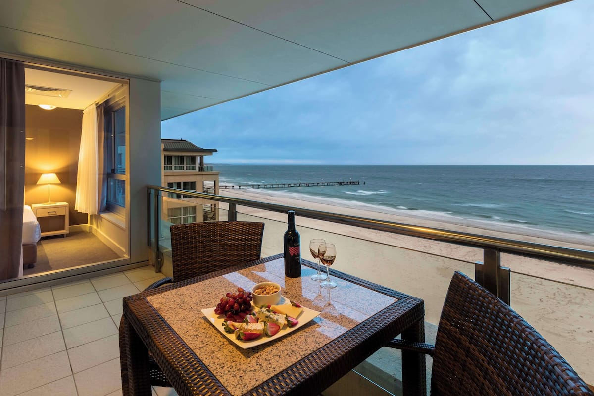Een balkon met een gedekte tafel, een fles wijn en een wijnglas. In de verte is de oceaan en een steiger te zien.