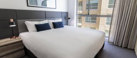 A hotel room with a large bed, a nightstand, a bedside lamp, a window with curtains, and a painting on the wall.