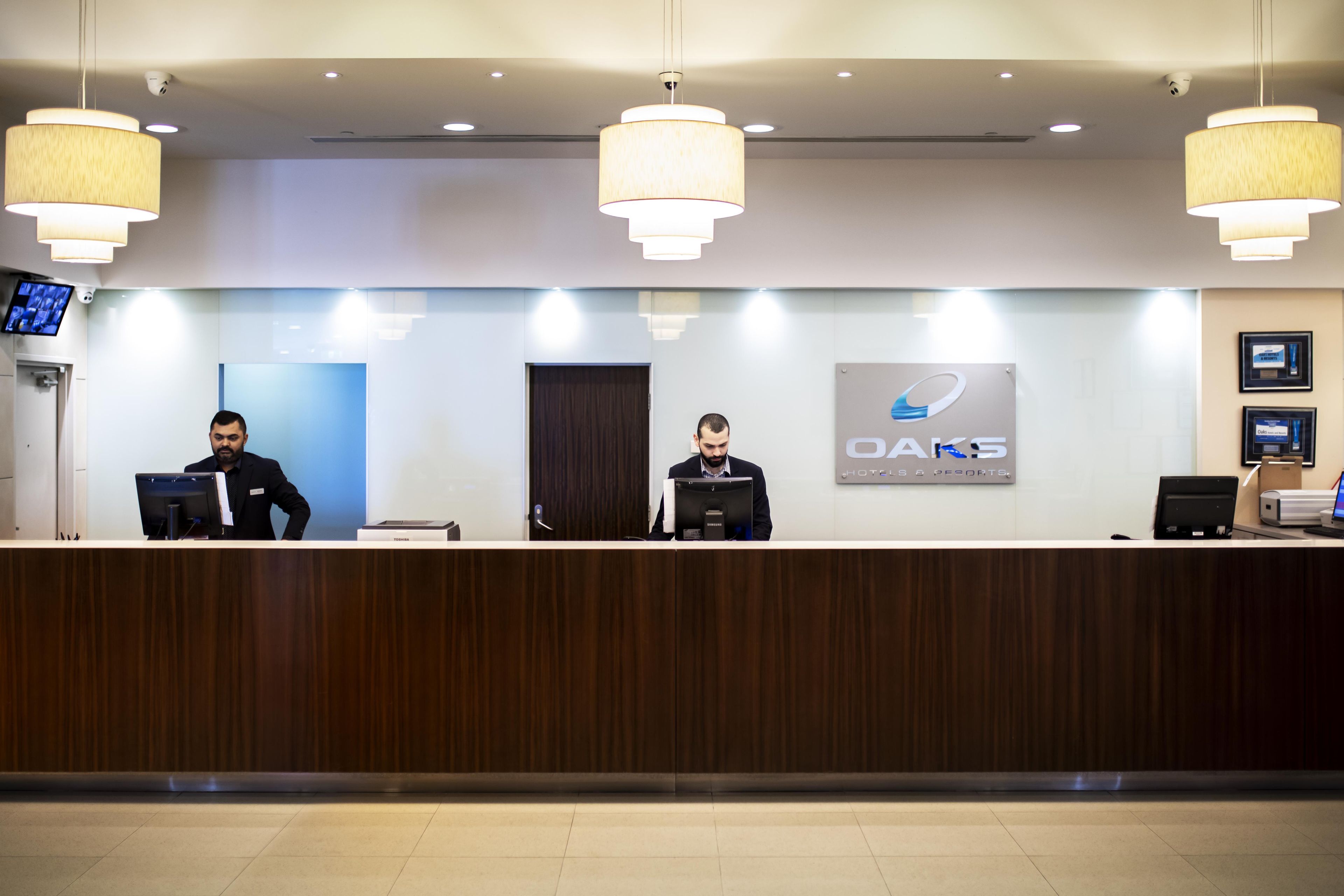 Una recepción con dos empleados, una pantalla digital y un letrero de OAKS Hotels & Resorts.