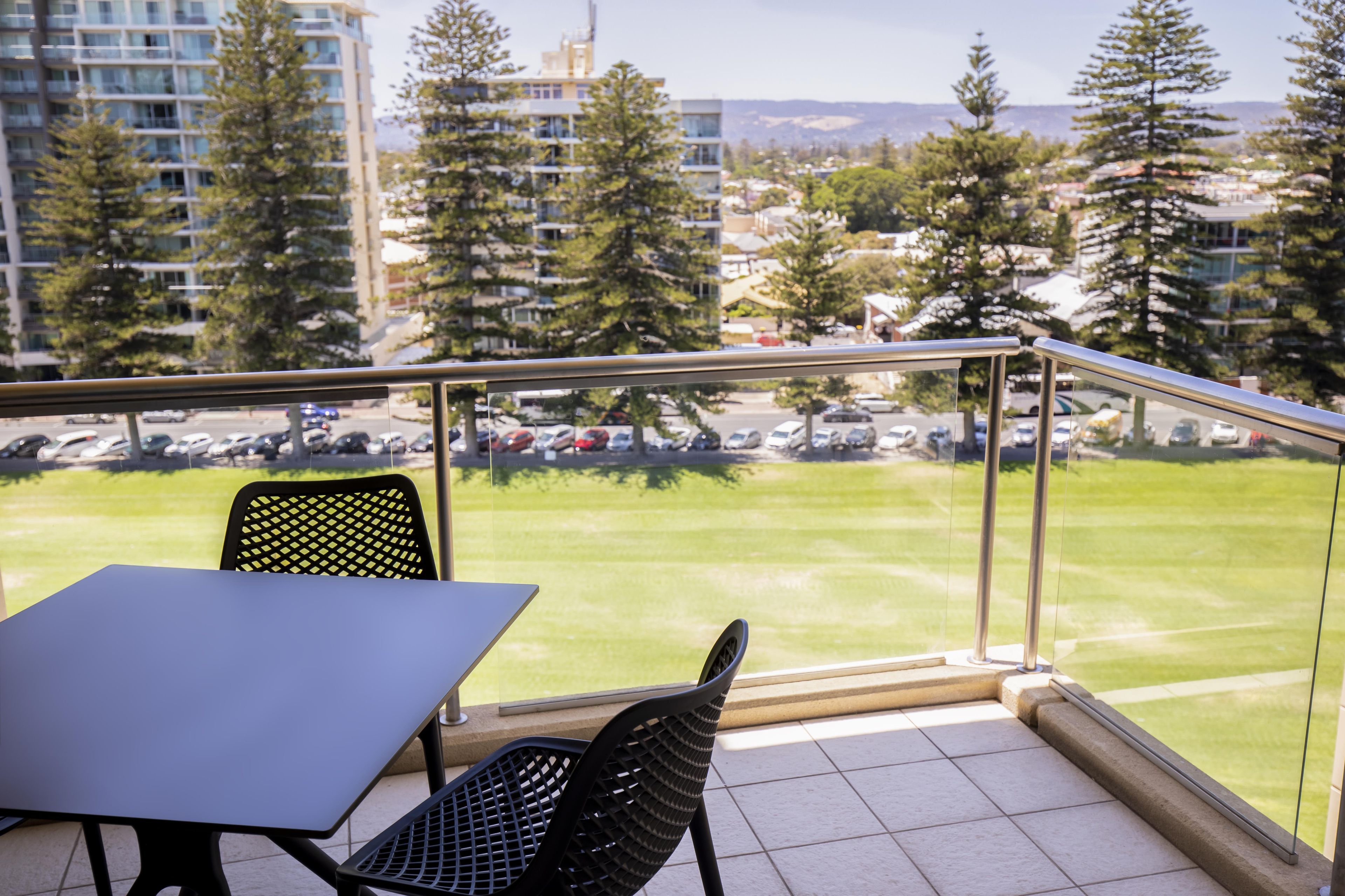 Un balcon avec une table et des chaises, donnant sur un parking et un champ verdoyant parsemé d’arbres.
