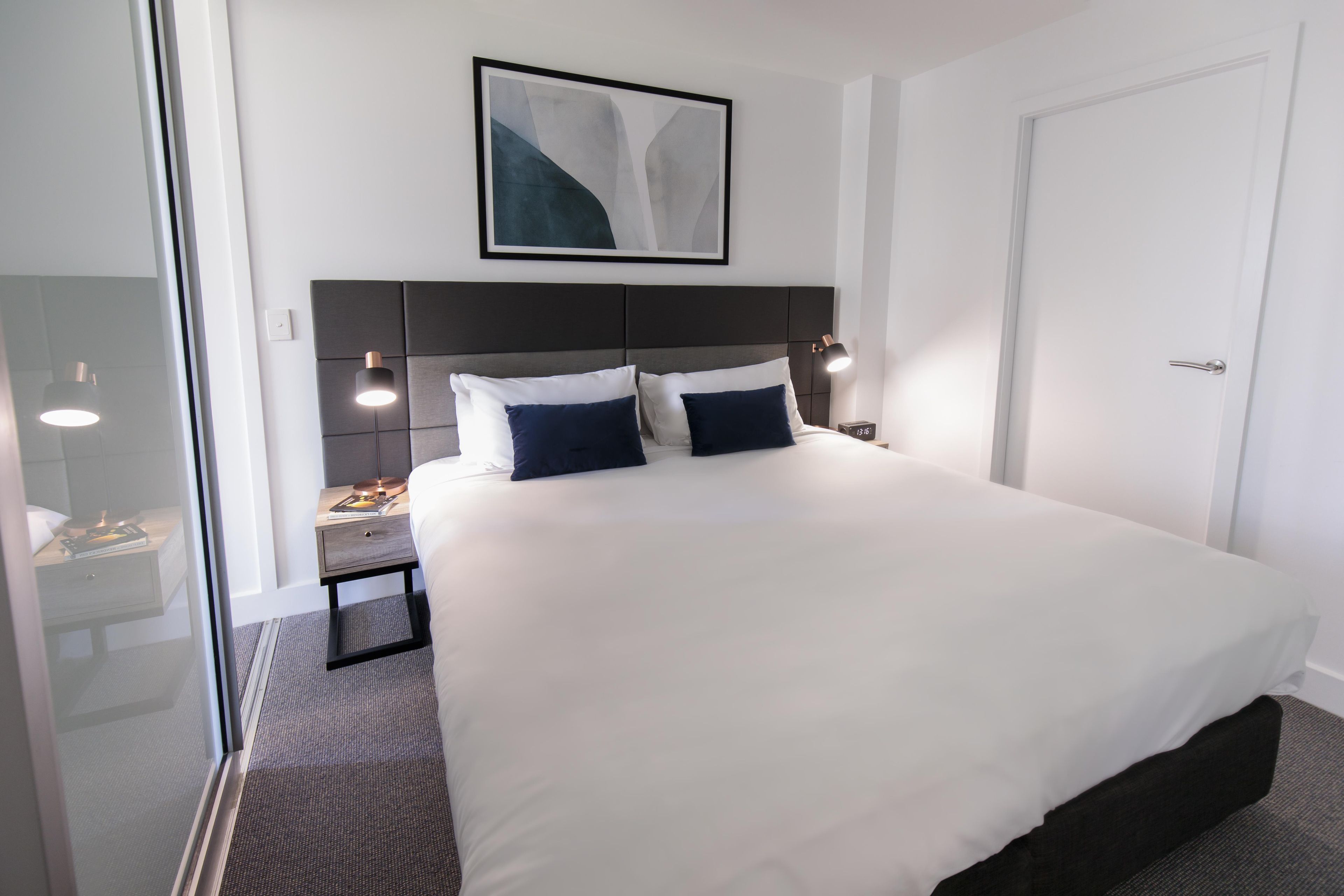 A hotel room with a large bed, a nightstand, a bedside table, a framed picture on the wall, and a door.