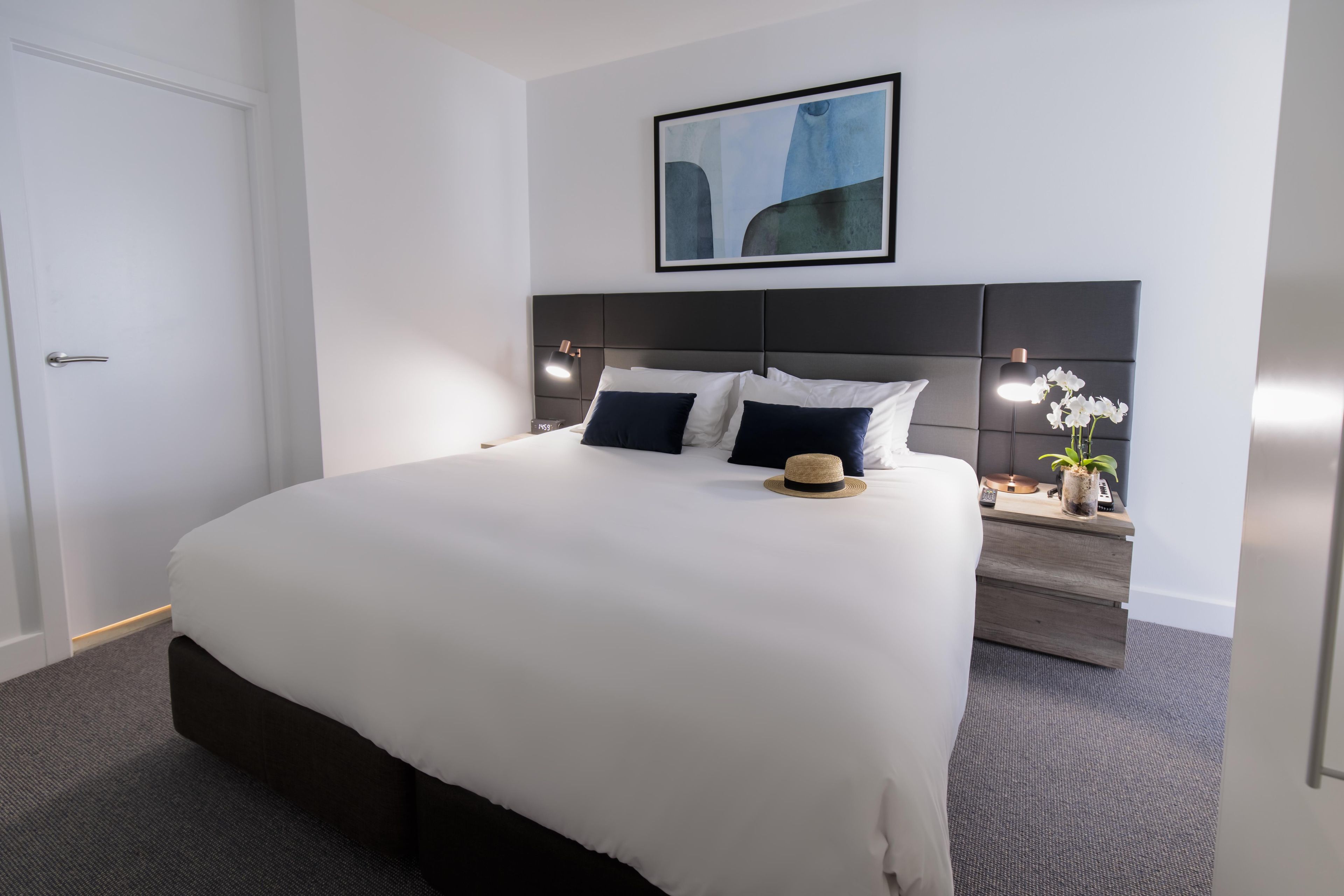 A hotel room with a large bed, a headboard, a bedside table with a lamp, and a framed picture on the wall.