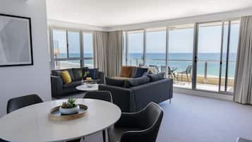 Una sala de estar moderna con un amplio ventanal con vistas a una playa, una mesa de comedor redonda y un sofá con cojines.