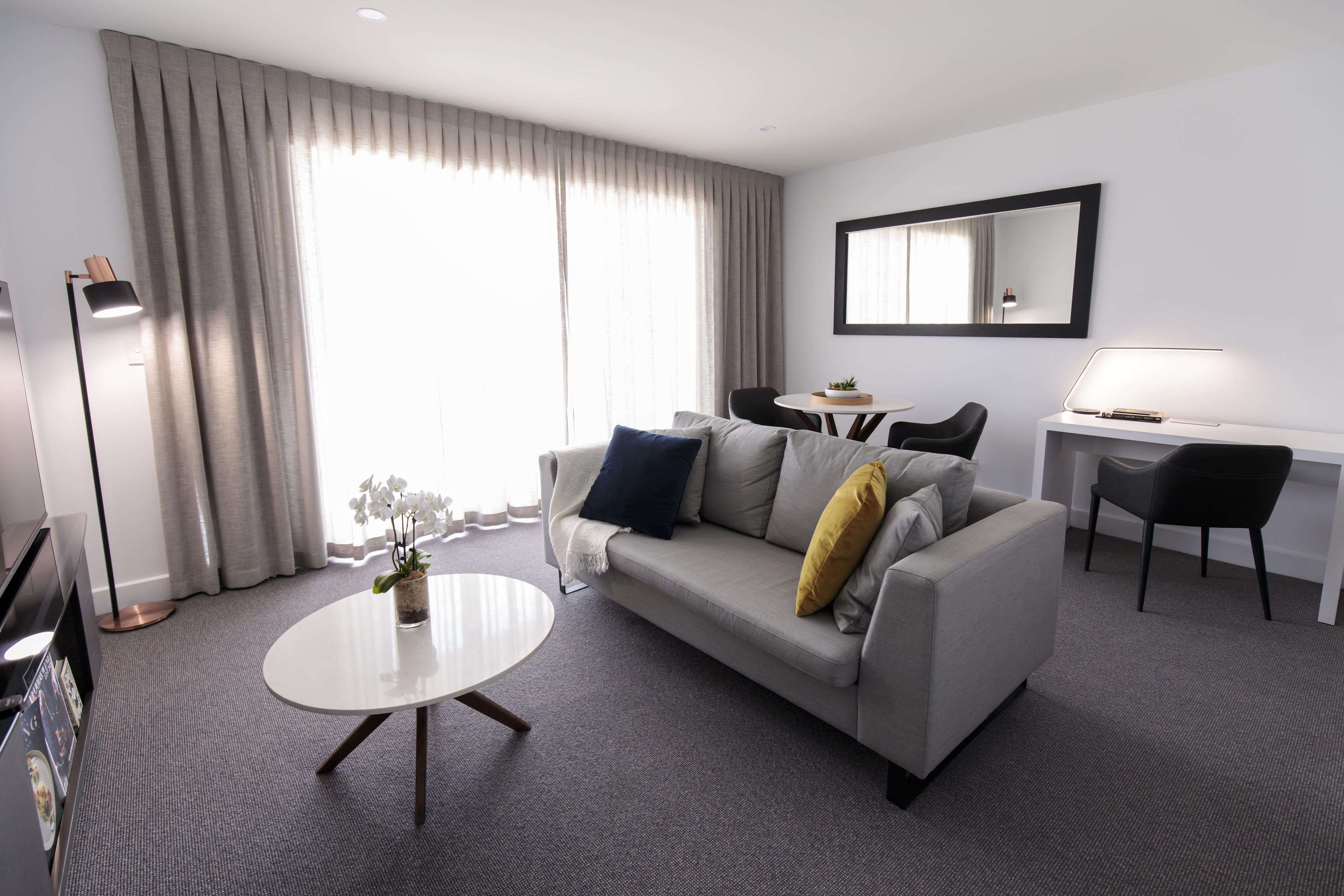 A modern hotel room with a grey sofa, a round coffee table, a desk with a chair, and a large window with curtains.