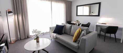 A modern hotel room with a grey sofa, a round coffee table, a desk with a chair, and a large window with curtains.