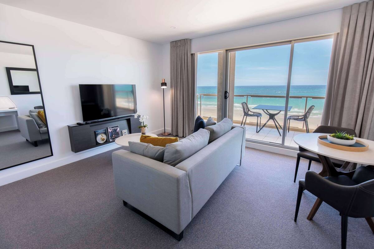Een moderne hotelkamer met een grijze bank, een flatscreen televisie, een zithoek met tafel en stoelen, en een balkon met uitzicht op de oceaan.
