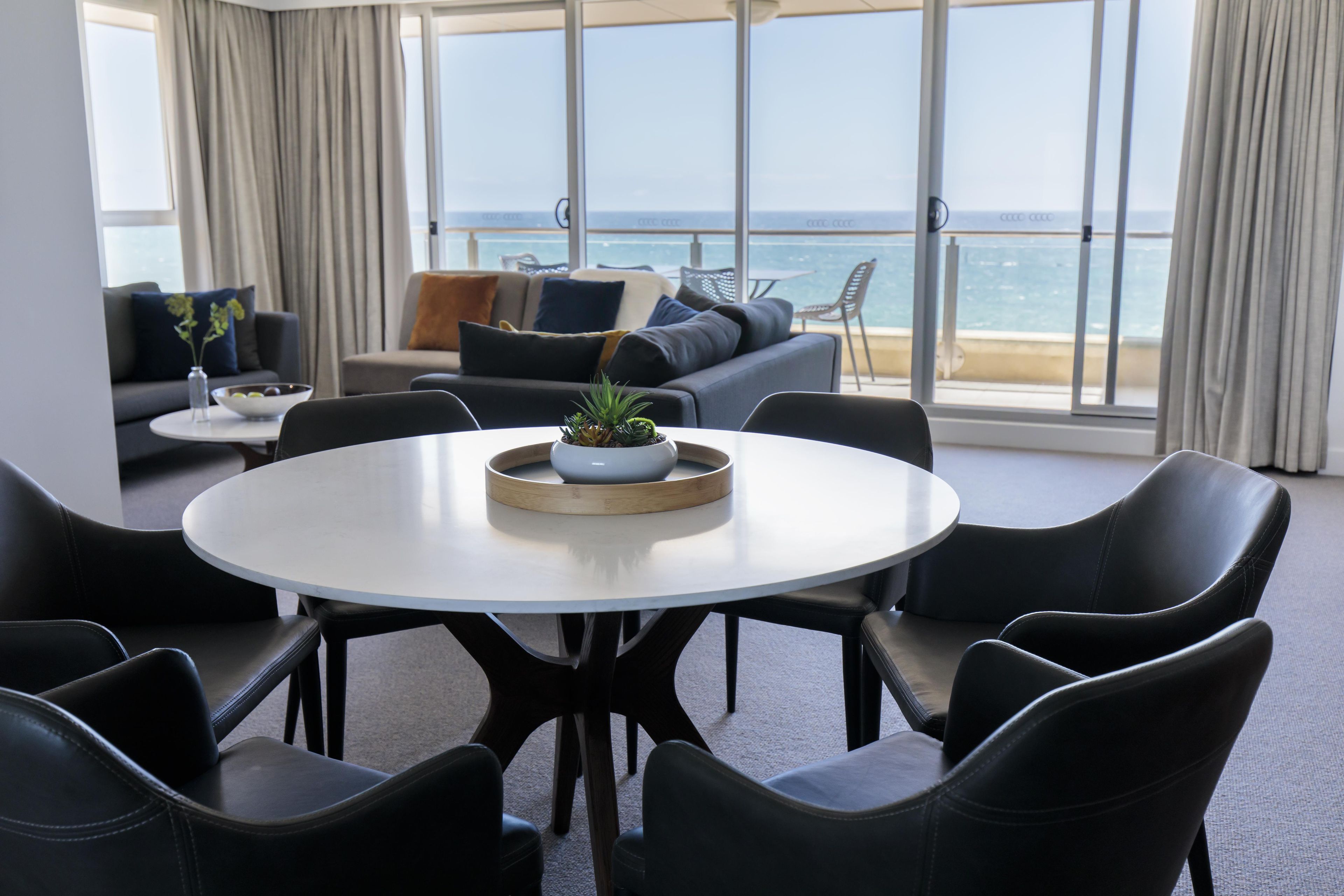 Ein modernes Wohnzimmer mit einem runden Esstisch, schwarzen Stühlen, einer Couch und Meerblick.