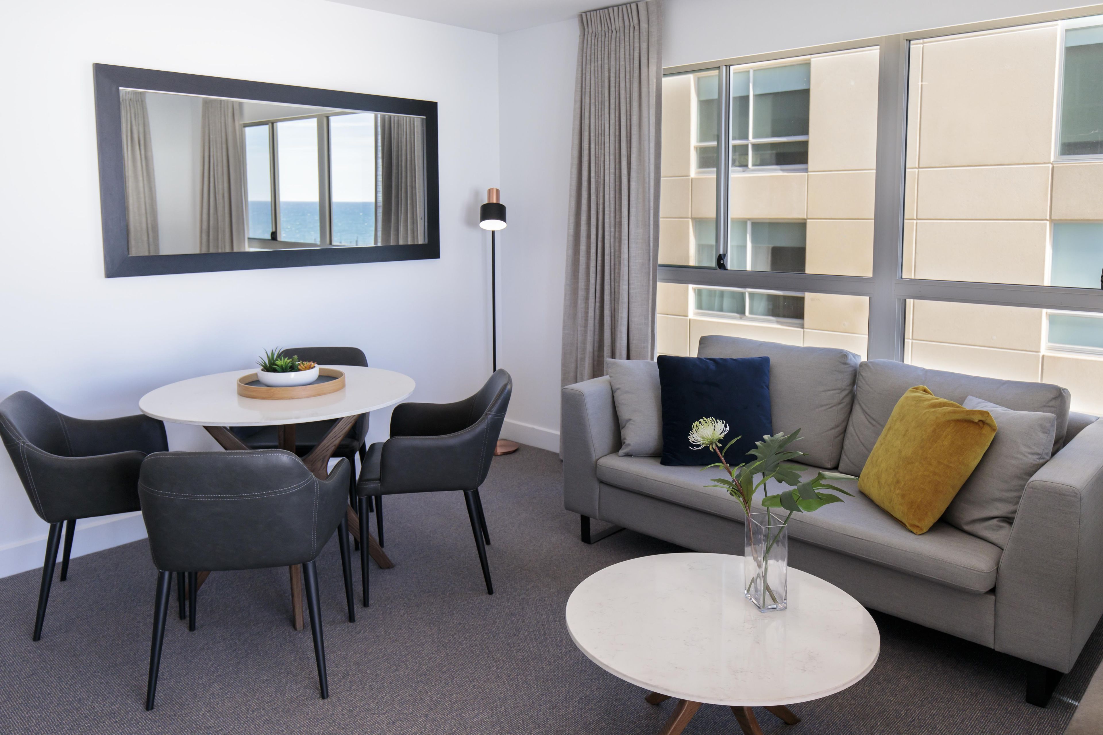 Un salon moderne avec une table de salle à manger ronde, deux chaises, un canapé avec des coussins, un lampadaire et une vue sur un immeuble à travers de grandes fenêtres.