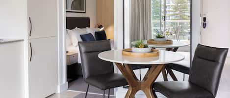 Habitación de hotel moderna con comedor que incluye una mesa redonda con sillas, una cama con cabecera y vistas al exterior a través de ventanas amplias.