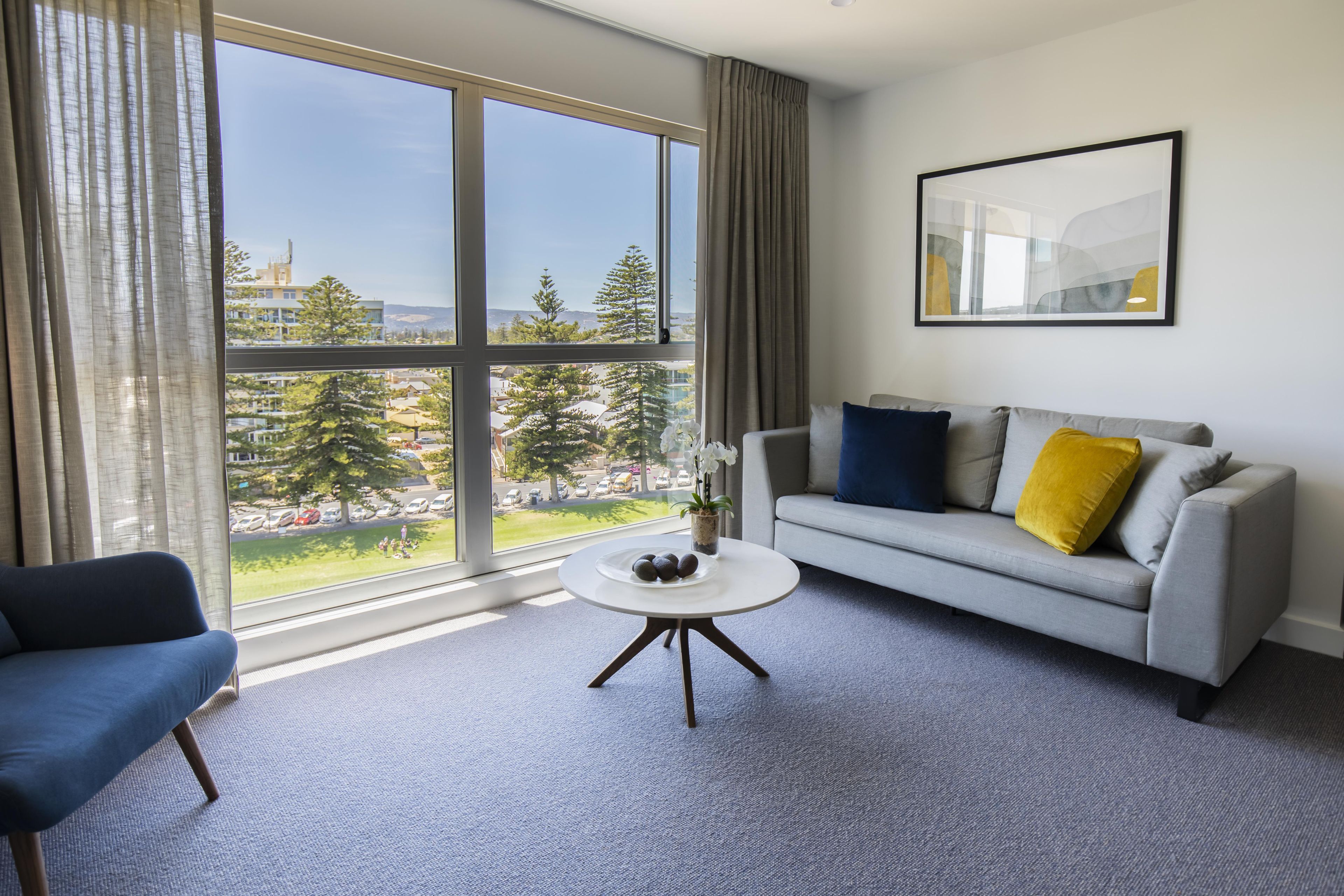 A modern hotel room with a large window, a grey sofa with yellow and blue cushions, a blue armchair, and a small white table.