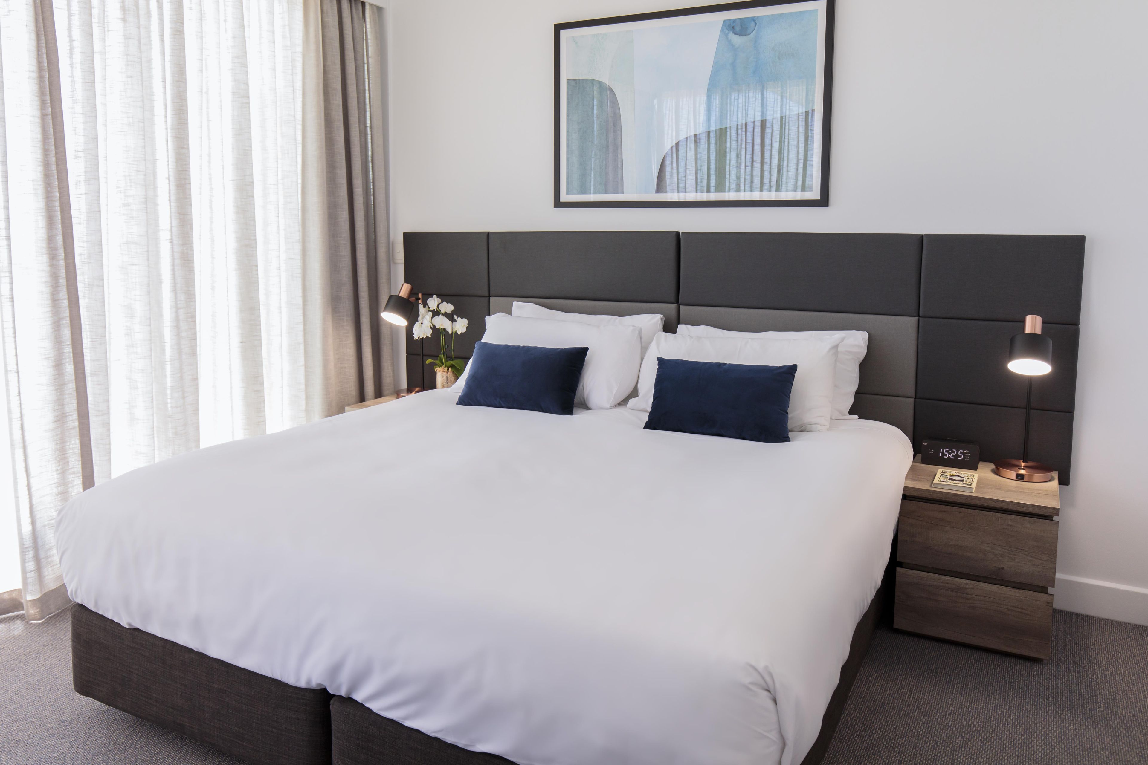 Une chambre d’hôtel avec un grand lit, deux tables de chevet, une petite table, une horloge et un tableau encadré au mur.