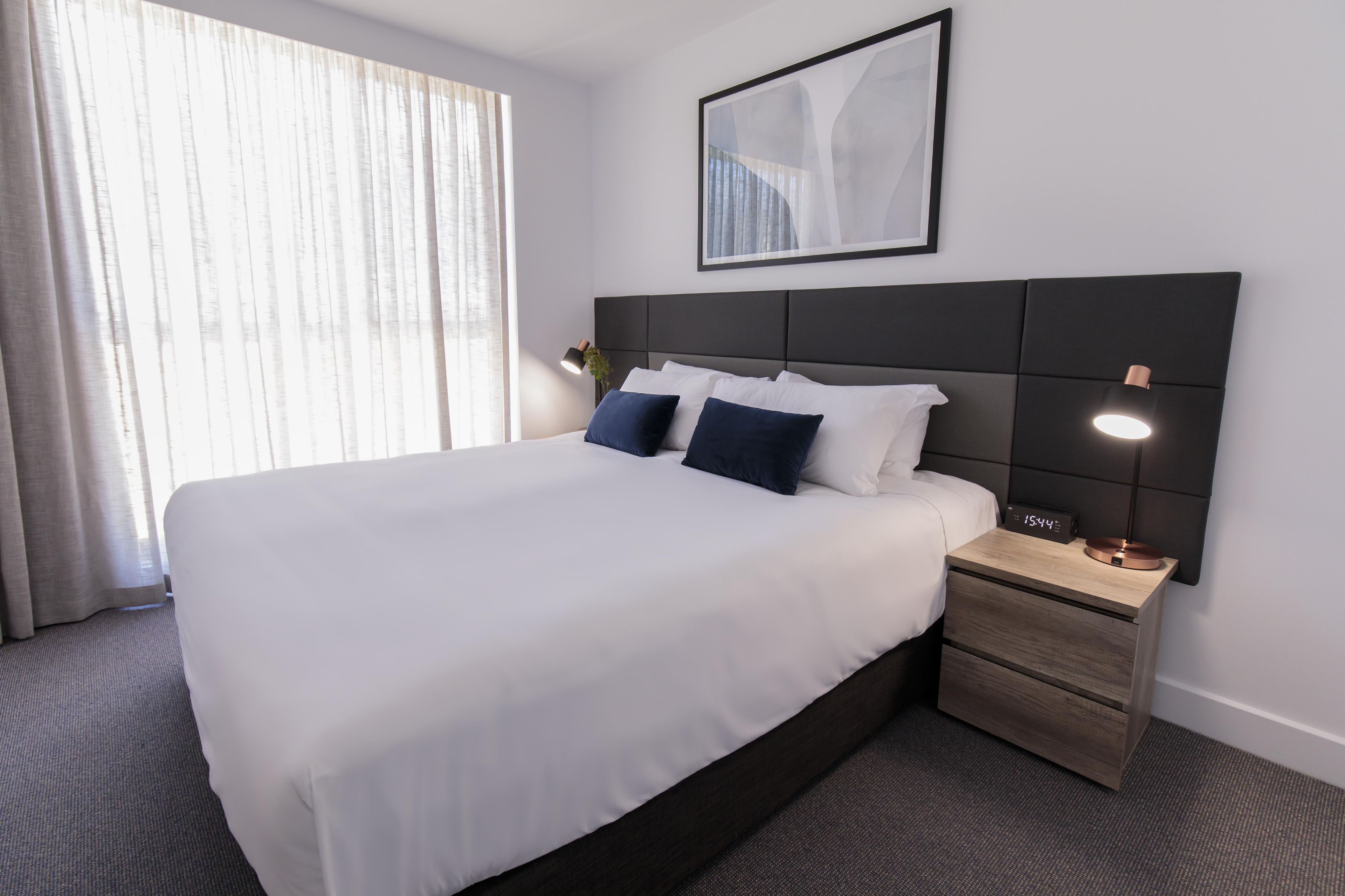 Une chambre d’hôtel avec un grand lit, une table de chevet avec une horloge et une fenêtre avec des rideaux.