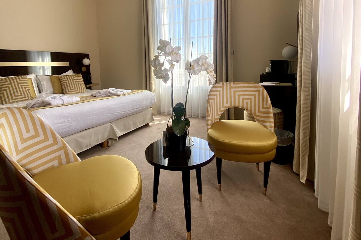 A hotel room with a bed, two armchairs, a small table, a desk, and a ceiling light fixture.