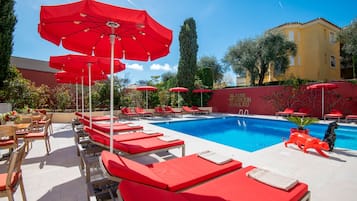 Un espace piscine avec des chaises longues rouges et des parasols, une piscine à l’eau d’un bleu limpide et un mur rouge orné d’un symbole de hashtag.
