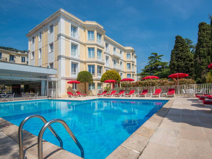 A swimming pool with red umbrellas and lounge chairs, surrounded by a building with multiple balconies.