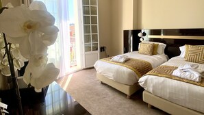 Ein Hotelzimmer mit zwei Betten, einem großen Fenster mit Vorhängen, einer Vase mit weißen Blumen und einem Schreibtisch mit Fernbedienung und einem Buch.