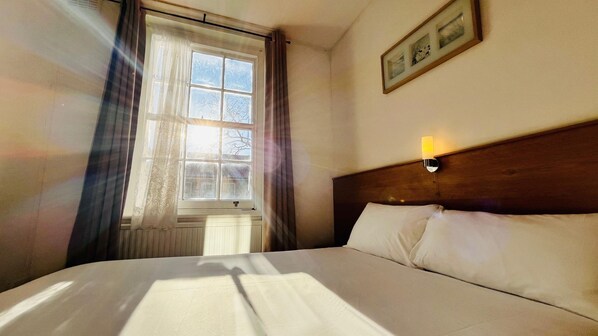 Una habitación de hotel con cama, ventana con cortinas, luz empotrada en la pared y un cuadro enmarco.
