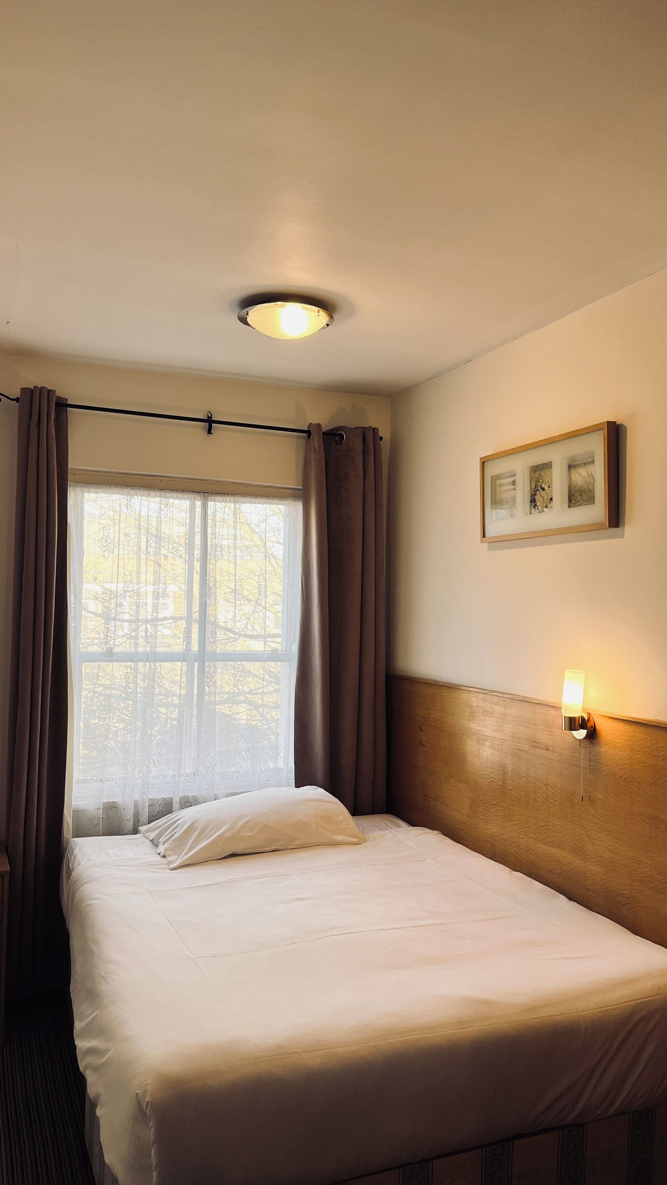 Ein Hotelzimmer mit einem Bett, einem Vorhangfenster, einer Wandleuchte und einem gerahmten Bild.