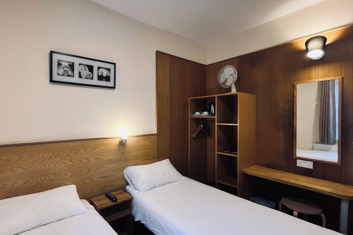 A hotel room with two single beds, a wooden wardrobe, a desk with a mirror, and a wall-mounted light.