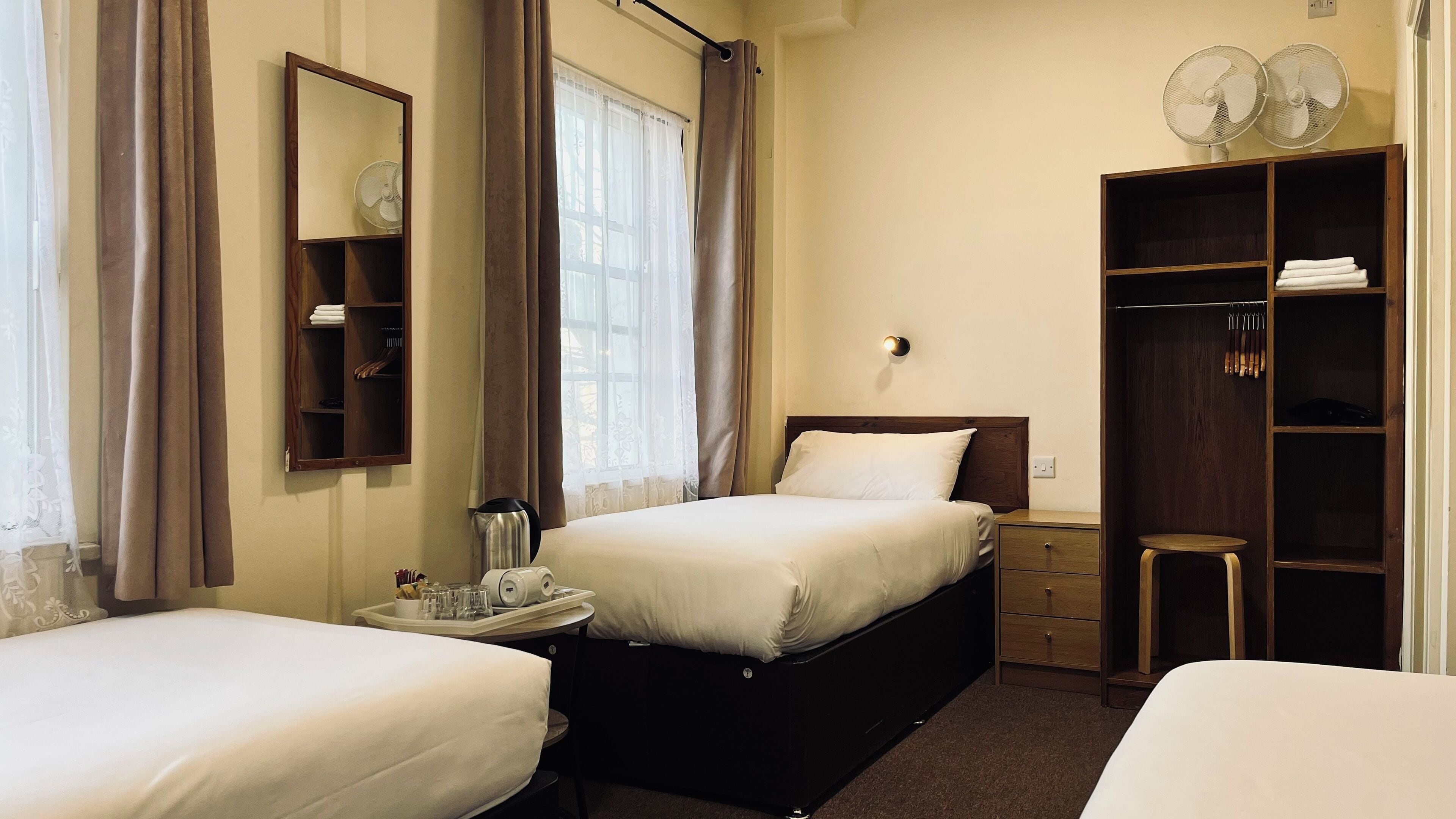 A hotel room with two beds, a bedside table with a tray of toiletries, a wardrobe, and a mirror on the wall.