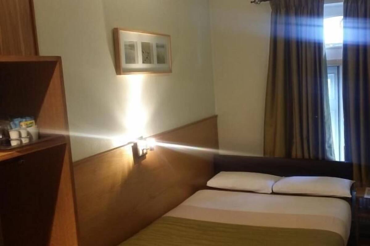 A hotel room with a bed, a bedside table with a lamp, a framed picture on the wall, and a window with curtains.