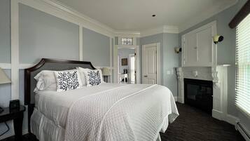 A bedroom with a large bed, a nightstand, a dresser, a mirror, a fireplace, and a door leading to another room.