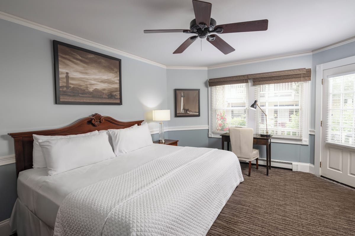 A hotel room with a large bed, a desk, a chair, a ceiling fan, and two windows with blinds.