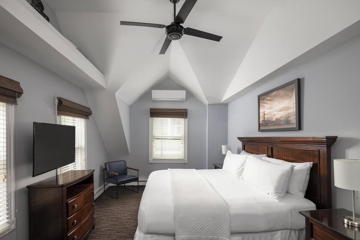 A hotel room with a bed, a TV, a desk, a chair, a ceiling fan, and a window with blinds.