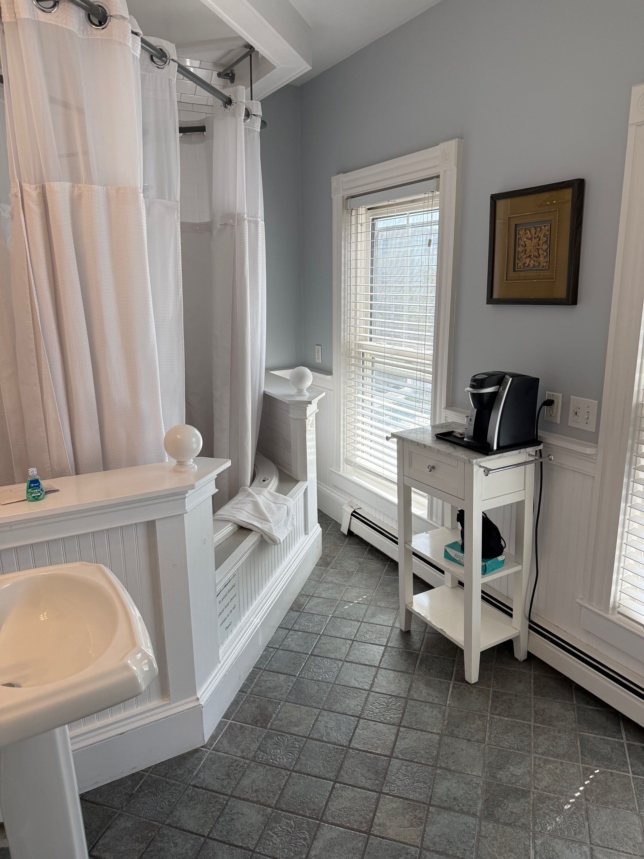 A bathroom with a white sink, a bathtub, a shower curtain, a window with blinds, a small white table with a coffee maker, and a framed picture on the wall.
