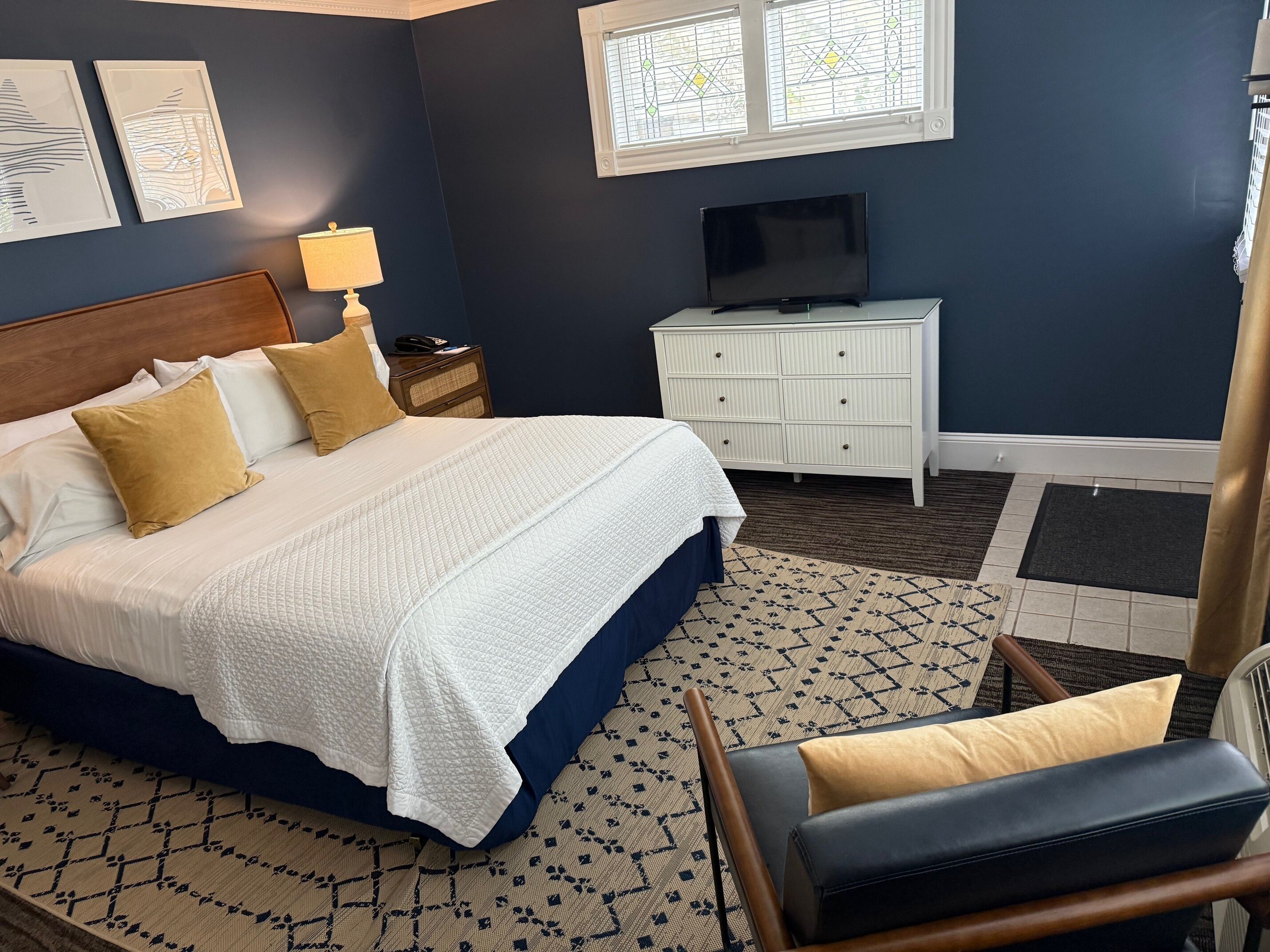 A neatly arranged bedroom with a bed, bedside table, lamp, TV, dresser, and patterned rug.