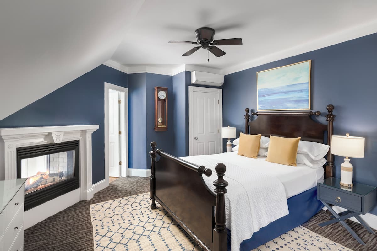 A bedroom with a bed, a nightstand, a lamp, a clock, a fireplace, and a ceiling fan.