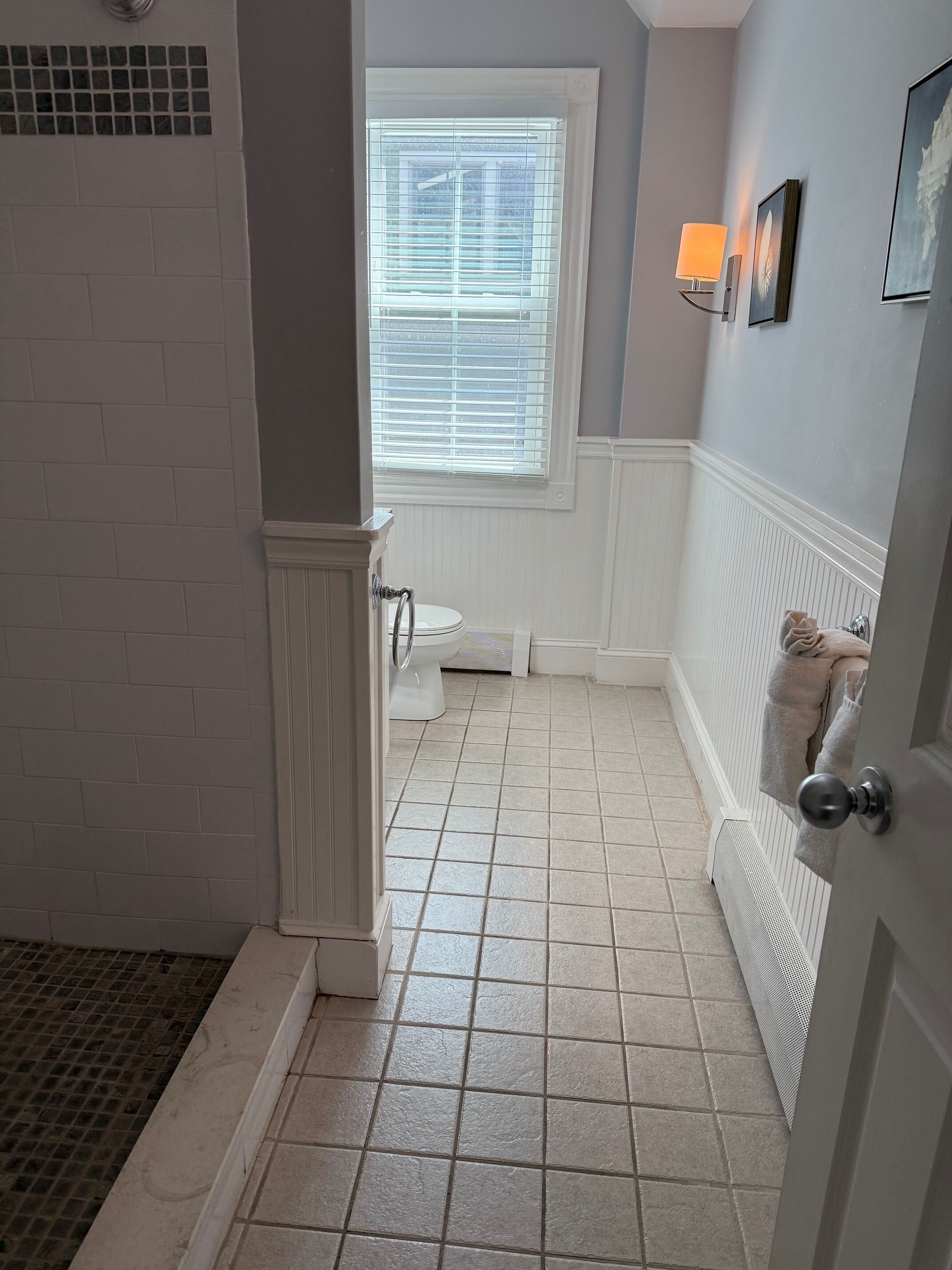 A bathroom with a toilet, a window with blinds, a towel rack with towels, and a door with a round knob.