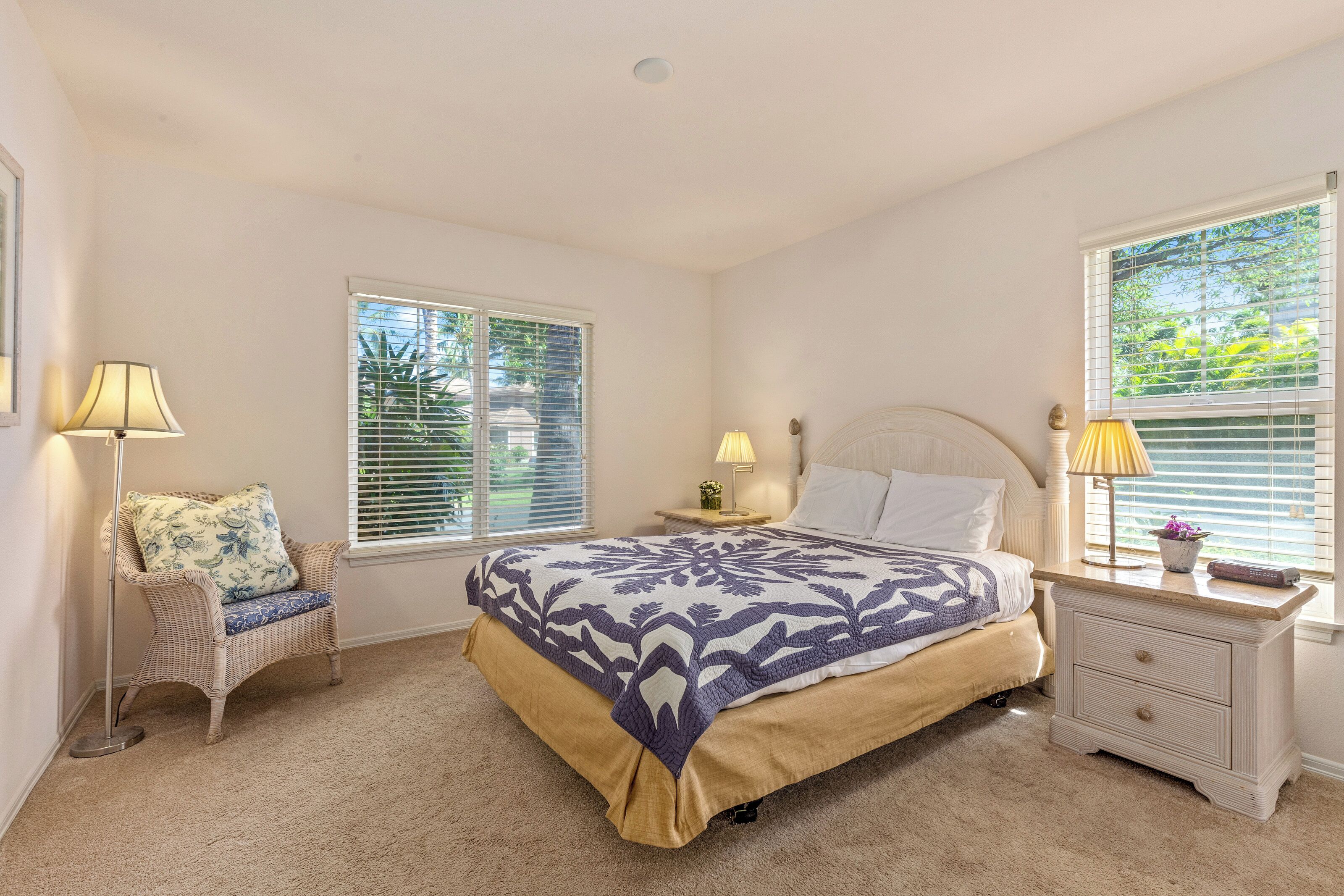 A bedroom with a bed, two bedside tables, a chair, and two windows with blinds.