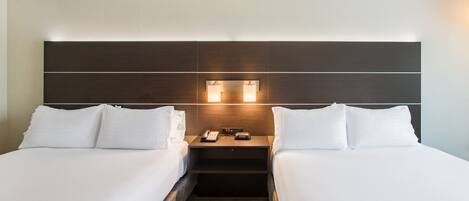 A hotel room with two beds, a nightstand with a phone and a clock, and a headboard with built-in lighting.