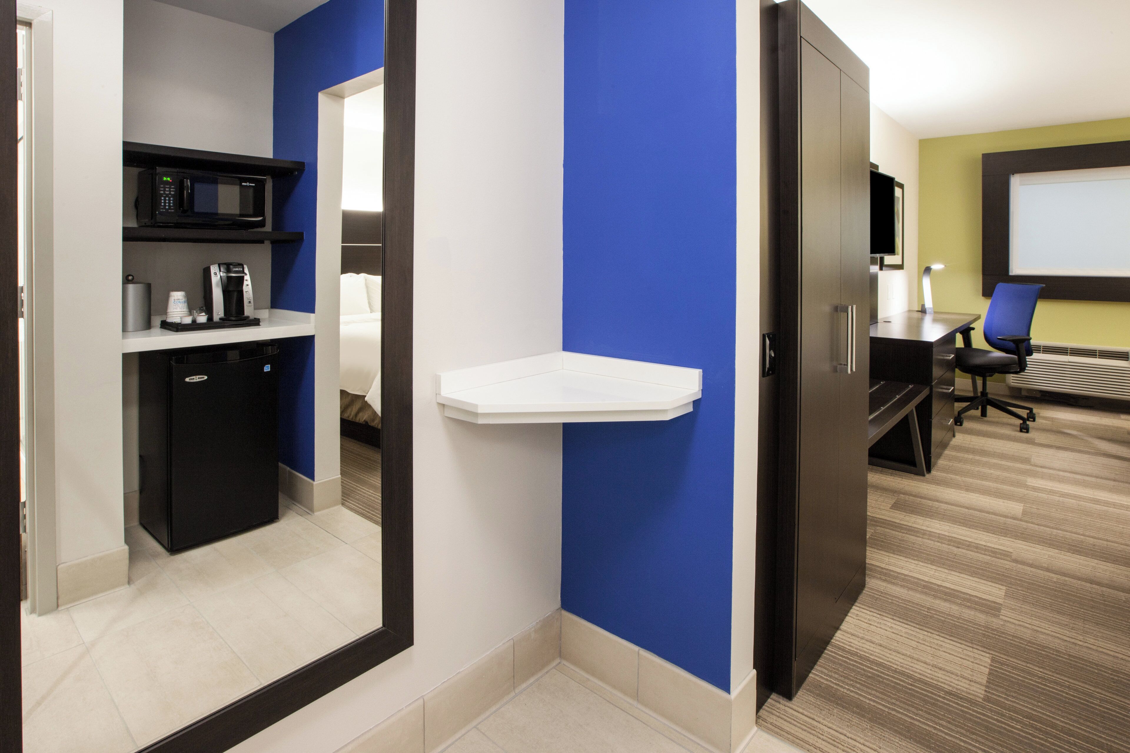 Een hotelkamer met een kitchenette, een wit schap, een bureau met stoel en een groot raam.