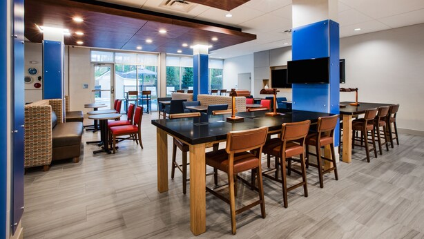 A modern dining area with multiple tables and chairs, a flat-screen TV mounted on the wall, and large windows providing natural light. - Holiday Inn Express Hotel & Suites Boston-Marlboro by IHG (Hudson)