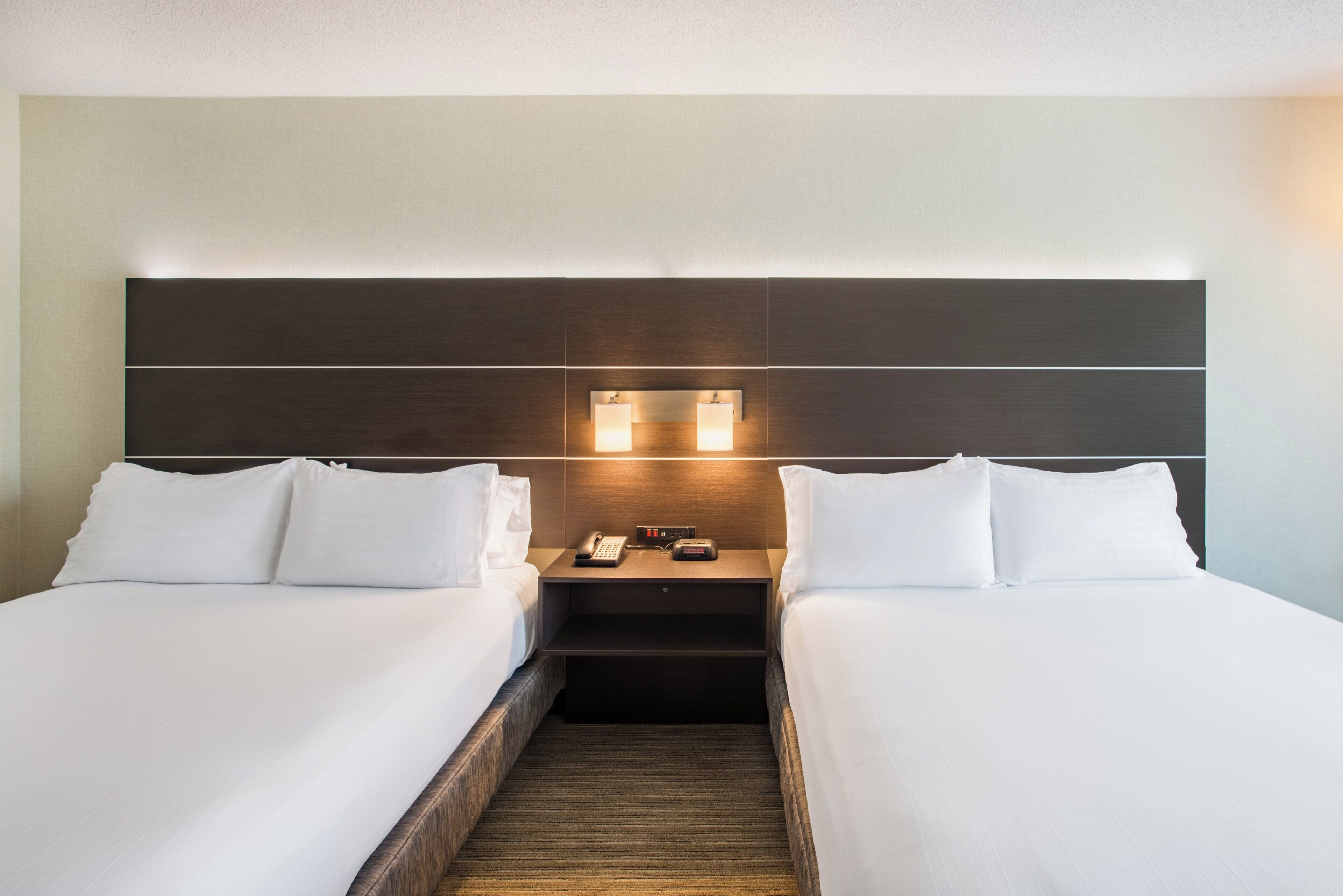 A hotel room with two single beds, a nightstand with a phone and a lamp, and a headboard with built-in lighting.
