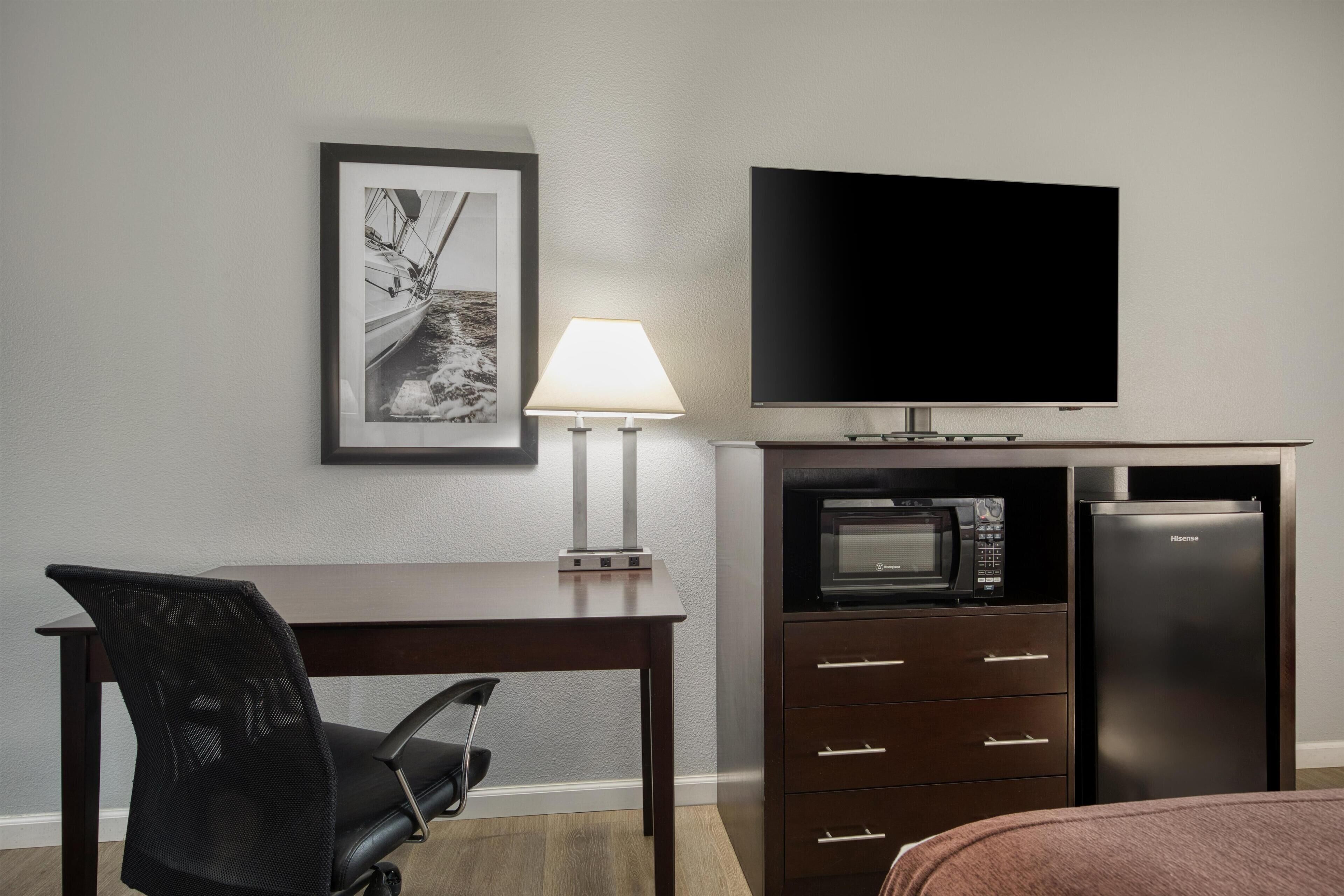 A hotel room with a flat-screen TV, a microwave, a refrigerator, a desk with a chair, and a framed picture on the wall.