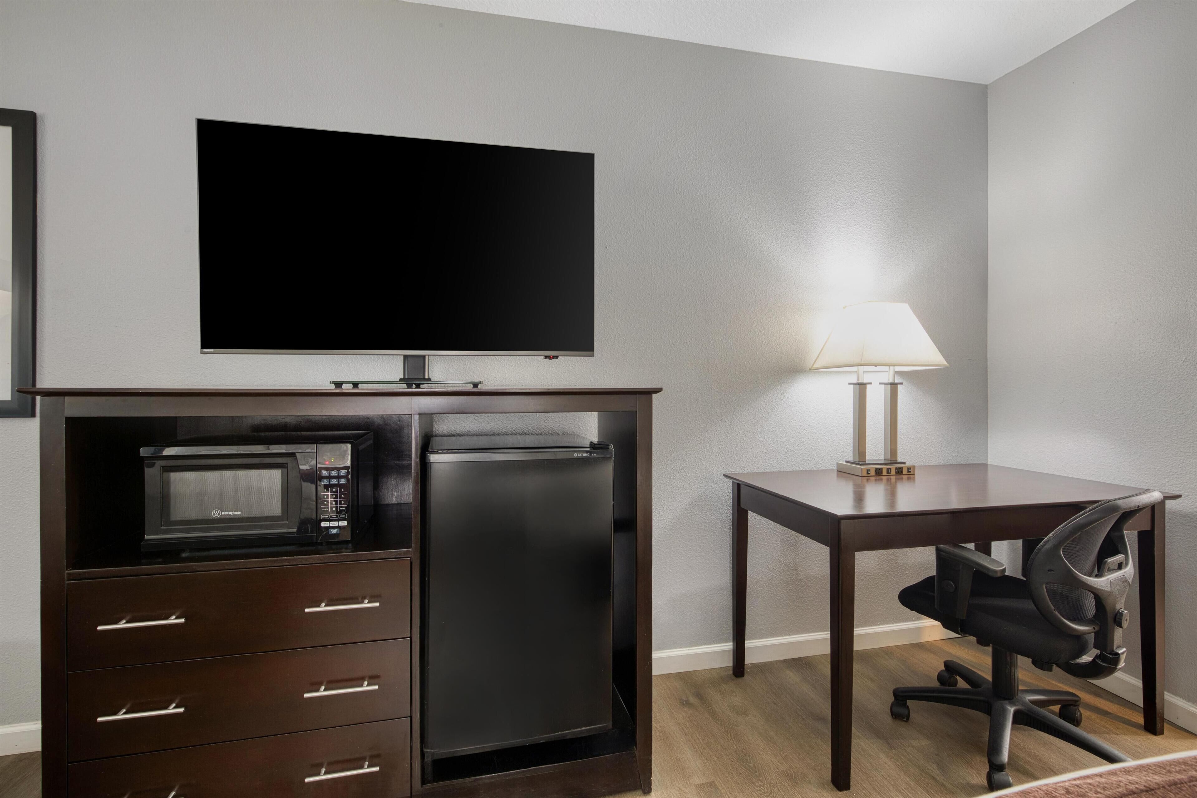 A compact hotel room with a flat-screen TV, a microwave, a mini-fridge, a desk with a chair, and a bedside lamp.
