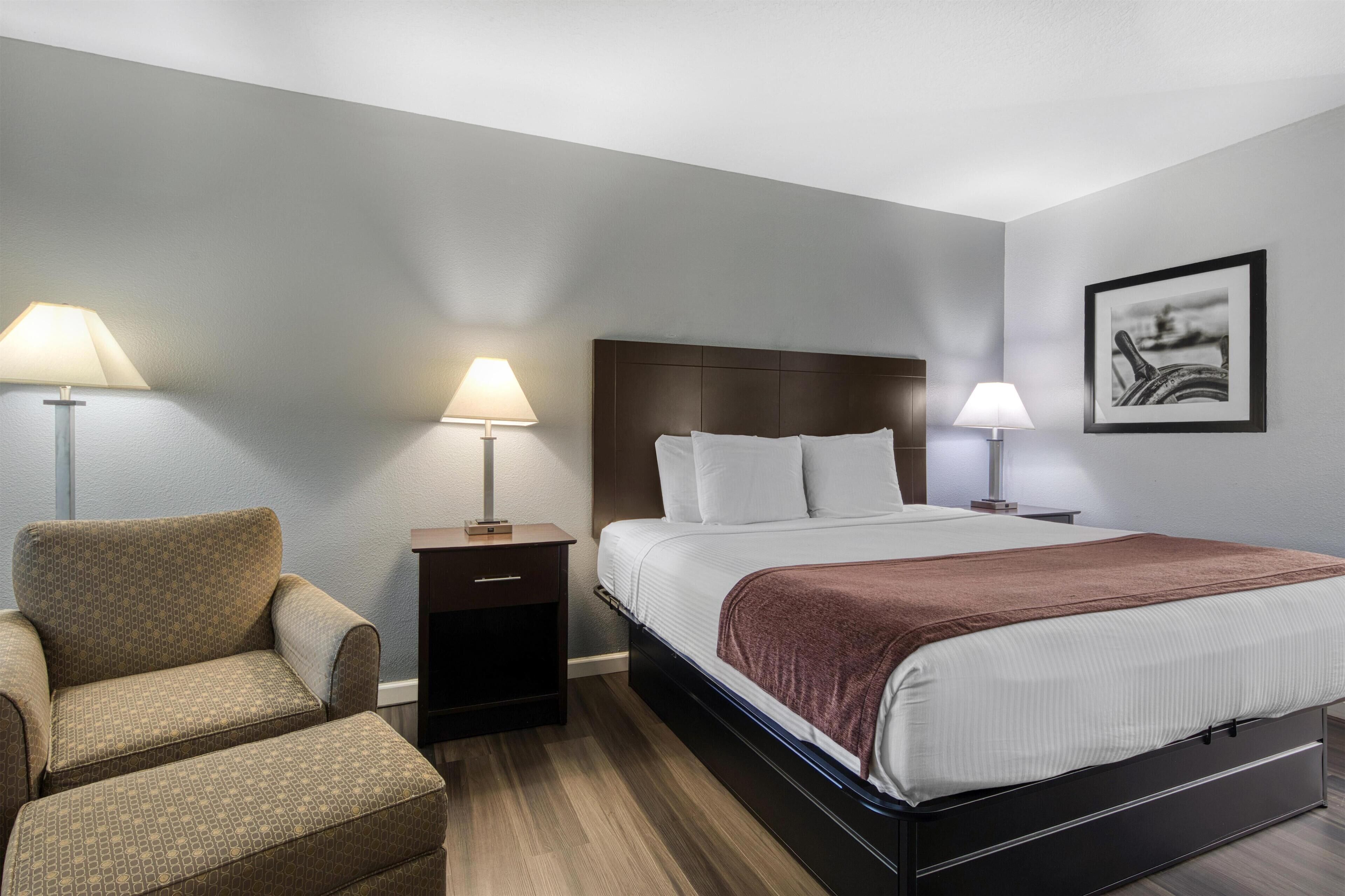 Une chambre d’hôtel avec un lit, deux lampes de chevet, une chaise, une table de chevet et un tableau encadré au mur.