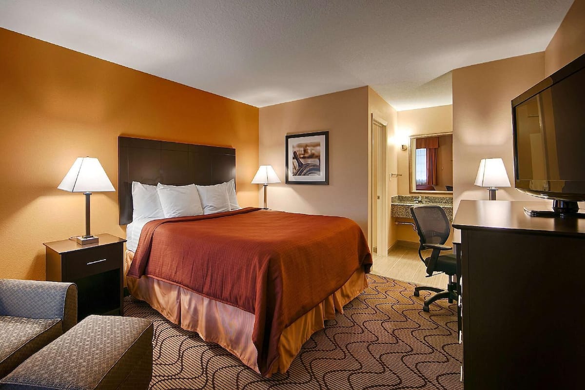 A hotel room with a bed, two bedside lamps, a desk with a TV, a chair, and a picture on the wall.