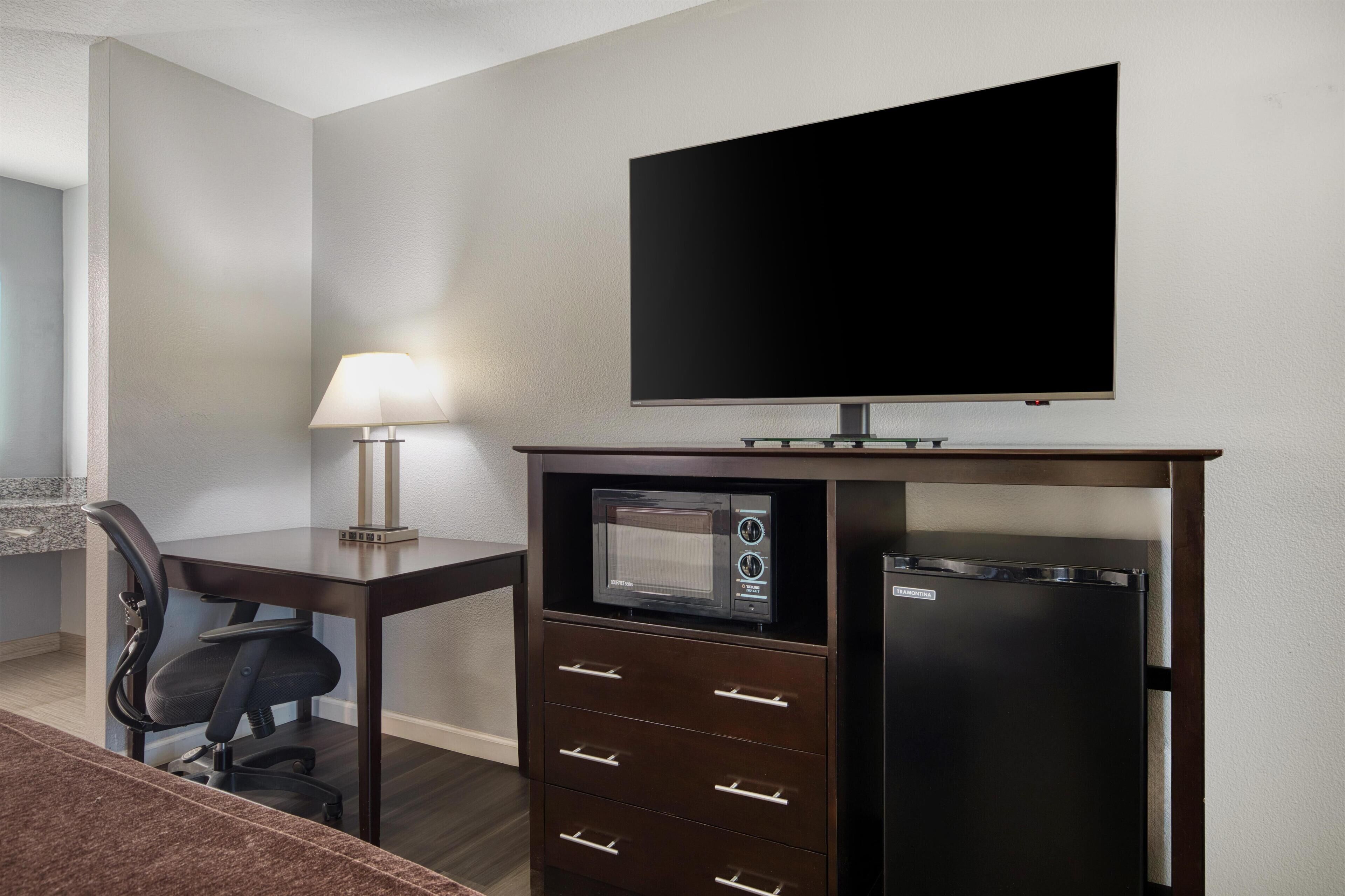 A hotel room with a flat-screen TV, a microwave, a refrigerator, a desk with a lamp, and a chair.
