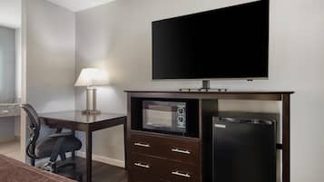 A hotel room with a flat-screen TV, a microwave, a refrigerator, a desk with a lamp, and a chair.