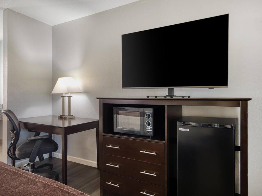 A hotel room with a flat-screen TV, a microwave, a refrigerator, a desk with a lamp, and a chair.