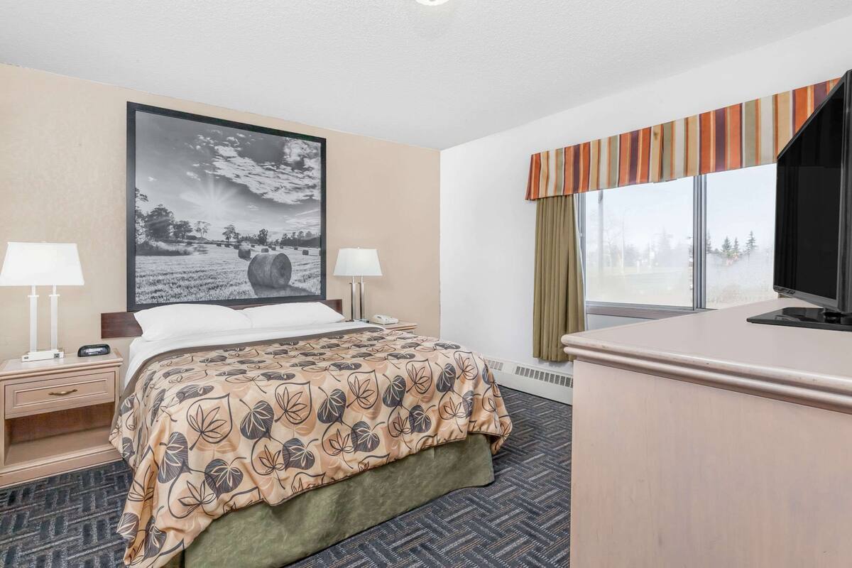 Hotelkamer met een bed, twee nachtkastjes met lampen, een groot ingelijst schilderij aan de muur, een bureau met een televisie, en een raam met gordijnen.