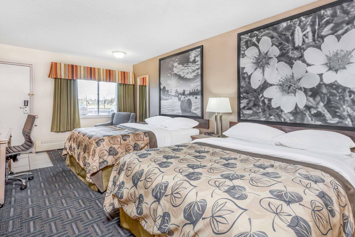 Hotelkamer met twee bedden, een bureau, een stoel, een raam met gordijnen en een groot, ingelijst kunstwerk aan de wand.