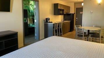 A compact hotel room with a bed, a kitchenette, a dining area with a table and chairs, and a door leading to a bathroom.