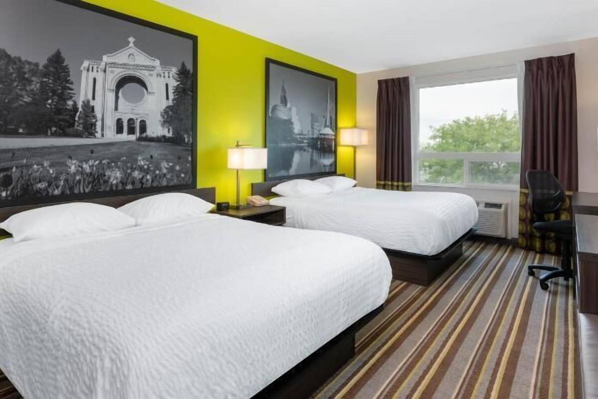 Habitación de hotel con dos camas, un escritorio, una silla, un ventanal grande y dos lámparas de pared.
