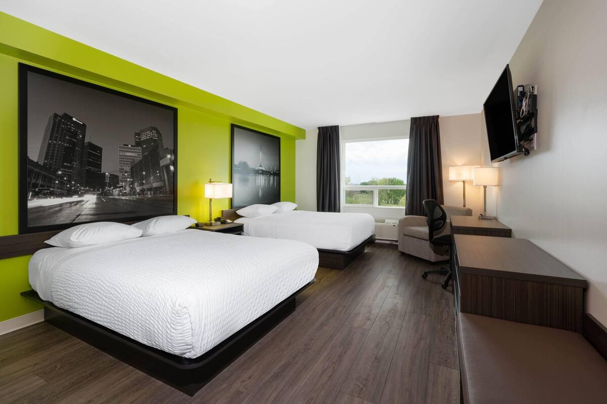 Habitación de hotel con dos camas, un escritorio, una silla, un televisor y un ventanal con vistas a la ciudad.